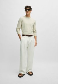 Helle beige Langarmpullover, weiße lockere Hose, brauner Gürtel und schwarze Flip-Flops. Accessoires umfassen übergroße schwarze Sonnenbrillen.