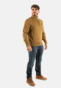 Sudadera marrón con cremallera y cuello alto, puños y dobladillo de canalé, combinada con jeans azul oscuro y botas de tobillo color beige.