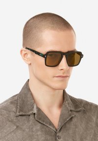 Ruskeansävyiset aurinkolasit, joissa on musta geometrinen kehys ja vihreämarmoroidut sankat. Suunnittelussa on ainutlaatuiset kulmikkaat reunat ja teksturoitu pinta.