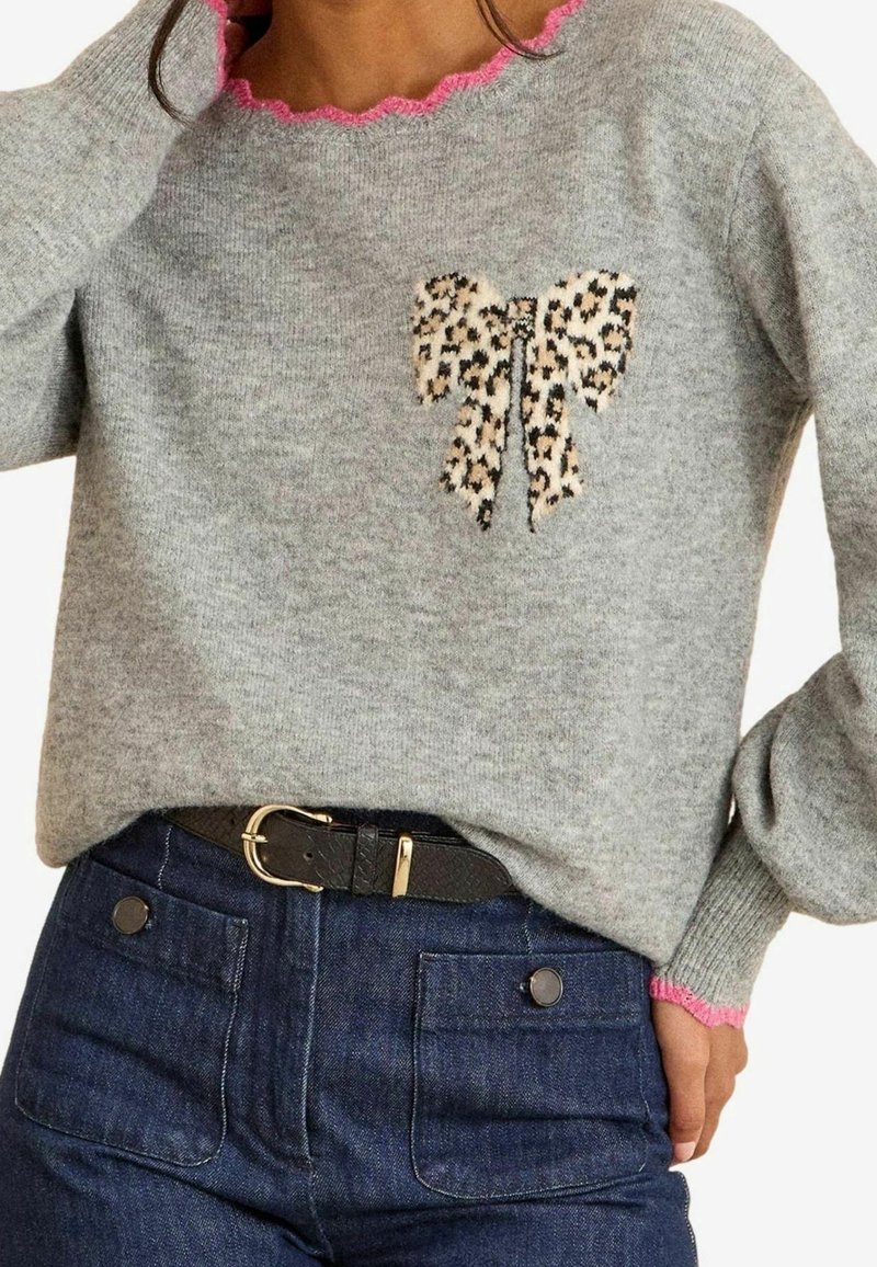 Pull gris avec bordures roses festonnées, motif nœud léopard sur la poitrine, associé à un jean bleu foncé et une ceinture noire avec boucle dorée.
