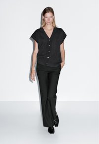 Frau, die geht, trägt ein schwarzes kurzärmliges, geknöpftes Oberteil, schwarze weite Hosen und schwarze Schuhe, vor einem schlichten weißen Hintergrund.