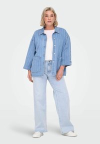 Gai zila džinsa jaka ar vertikālām svītrām, lieliem pogām un divām kabatām; apvienota ar gaiši ziliem platām kājām džinsiem un baltiem sporta apaviem.