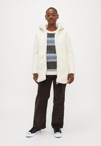 Veste en polaire blanche et moelleuse avec capuche, portée sur un pulls rayé aux tons bleu et noir, associée à un pantalon en velours côtelé marron et des baskets noires.
