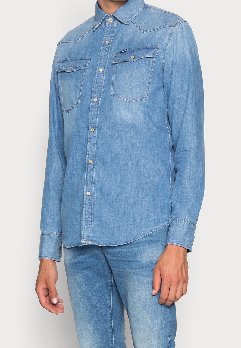 Ljusblå denimsjort med långa ärmar, tryckknappar, två bröstfickor, spetsig krage och rundad fåll, tillverkad av mjukt bomullsmaterial.