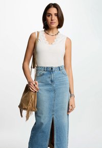 Top blanco sin mangas con detalles de encaje, combinado con una falda de mezclilla de tiro alto que presenta una abertura frontal. Bolsa marrón con flecos y joyas visibles.