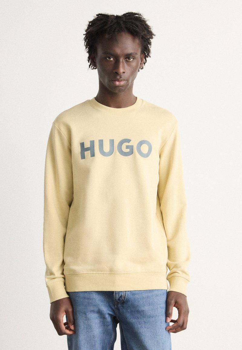 Lys gul hettegenser med "HUGO" trykt i grått, med rund hals og lange ermer, brukt sammen med blå jeans. Slette tekstur.