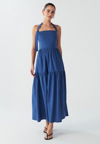 Modré maxi šaty s priliehavým živôtikom a ramienkami na zaväzovanie za krk, s viacvrstvovou sukňou a jemnou textúrou. Doplněné detailom v podobe mašle na boku.