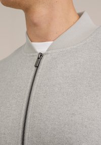 Světle šedá bomberová bunda s herringbone texturou, vybavená předním zipem a žebrovaným límcem. Hladká látka s minimalistickým designem.