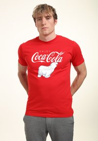 Rød bomulds t-shirt med teksten "Enjoy Coca-Cola" og et hvidt isbjørne grafik på fronten. Korte ærmer og rund hals design.