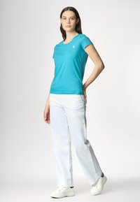 Una donna sta in piedi con sicurezza indossando una maglietta turchese brillante e pantaloni bianchi, con scarpe da ginnastica bianche, su uno sfondo grigio chiaro.