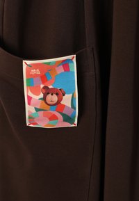 Brązowa tkanina zdobi duża metka z kolorowym abstrakcyjnym wzorem i ilustracją niedźwiedzia, obszyta czerwonym ściegiem.