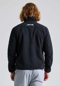 Schwarze Fleecejacke mit hohem Kragen, ausgestattet mit einem schlanken Design und dezenten Steppdetails. Modell von hinten betrachtet.