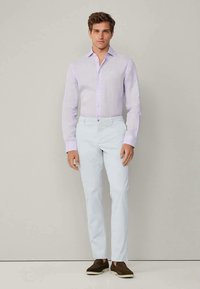 Man wearing light purple long-sleeve button-up shirt, white pants, and dark brown slip-on shoes standing against a plain background.