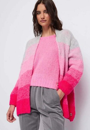 Donna con maglione rosa a maglia e cardigan aperto sfumato dal rosa al grigio, abbinato a pantaloni grigi plissettati, in piedi davanti a uno sfondo bianco.