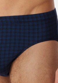 Dunkelblaue Badebriefe mit einem hellblauen Karomuster. Glattes Material mit enganliegendem Schnitt und elastischem Bund. Ein Bein ist teilweise sichtbar.