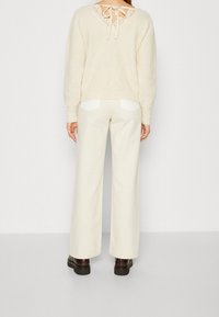 Off-white trui met een met kant afgewerkt decolleté en strik op de rug, gecombineerd met crèmekleurige wijde broek met witte accentzakken.