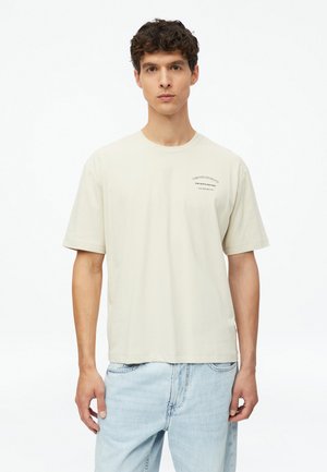 Young man with short curly hair wearing a light beige T-shirt with small black text on left chest and light blue jeans against white background.