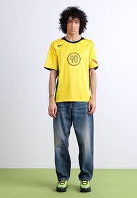 Yellow Nike jersey with black accents and a circular '90' logo at the front, paired with loose blue denim jeans and green-accented sneakers.