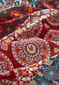 Tissu avec des motifs floraux et géométriques colorés et complexes en rouge, bleu, orange et blanc, présenté en plis doux et en vue rapprochée.