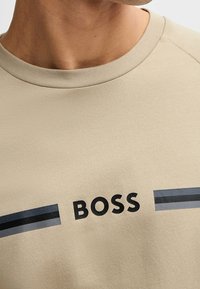 Sweatshirt beige à col rond avec une finition texturée, présentant des rayures horizontales noires et bleues. Logo "BOSS" imprimé en noir gras.