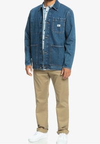 Kurtka dżinsowa w ciemnym granacie, z dwoma kieszeniami na piersi, zapinana na guziki, z kontrastującym ściegiem. Nosi się ją na jasną koszulę w kratę i beżowe spodnie.