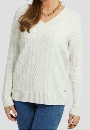 Woman wearing a white cable-knit V-neck sweater, blue jeans, gold chain necklace, and rings on left hand against plain background.