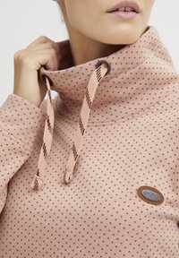 Pullover in pile rosa chiaro con motivo a pois bordeaux, colletto alto e cordini regolabili. Presenta un'etichetta in pelle. Texture morbida.
