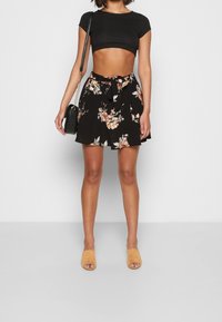 Jupe noire à motifs floraux avec une taille nouée, associée à un crop top noir ajusté. Escarpins beige et un petit sac bandoulière noir.