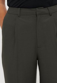 Personne portant un pantalon olive foncé sur mesure et une chemise noire, vue de la taille au milieu des cuisses, les mains dans les poches.
