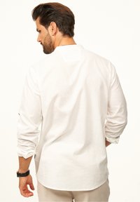 Weißes Leinenhemd mit langen Ärmeln, Rundhalsausschnitt und einer dezenten Textur. Verfügt über umgeschlagene Manschetten und einen abgerundeten Saum, von hinten betrachtet.