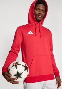 Κόκκινο φούτερ με μαλακό ύφασμα και μπροστινή τσέπη. Διαθέτει λευκό λογότυπο Adidas. Χέρια κρατούν μια μπάλα ποδοσφαίρου με ασπρόμαυρα αστέρια.