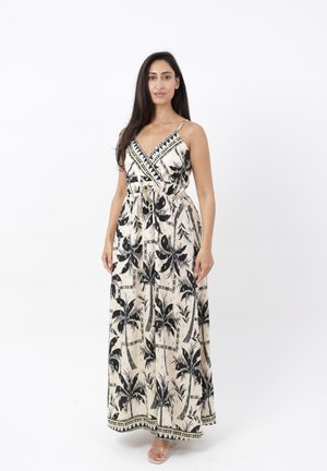 Woman wearing sleeveless, ankle-length beige dress with black palm tree prints, standing against a white background with hands by sides.