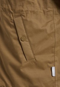 Tissu de veste imperméable marron en gros plan, avec une poche latérale munie d'un bouton rond et d'une étiquette détaillée. Texture lisse et finition mate.