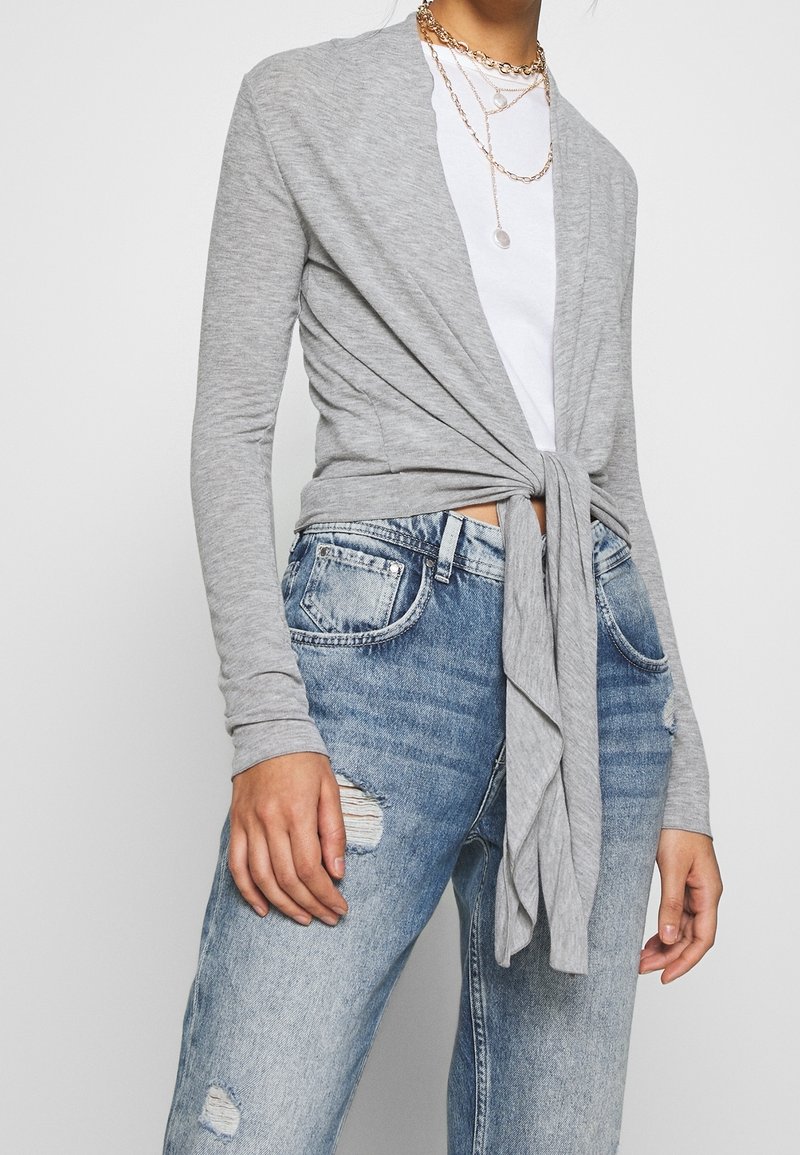 Grå mjukstickad cardigan med knyt framtill, bärs över en vit t-shirt och slitna blå jeans, med en avslappnad passform och långa ärmar.