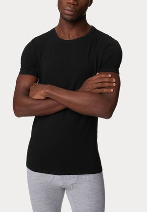 Schwarzes T-Shirt mit kurzen Ärmeln in taillierter Passform, aus glattem Stoff, getragen mit hellgrauen strukturierten Hosen. Das Modell posiert mit verschränkten Armen.