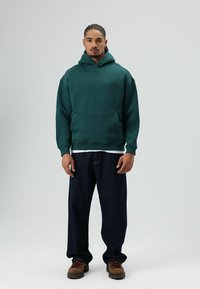 Sweat à capuche vert foncé avec une poche avant, porté sur une chemise blanche. Associé à un jean ample foncé et des chaussures marron à lacets.