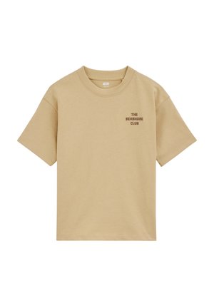 Beżowy t-shirt z krótkim rękawem i okrągłym dekoltem, z małym brązowym napisem "THE SEASHORE CLUB" na górnej lewej części klatki piersiowej.
