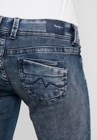 Jeans in denim blu scuro con zone sbiadite, caratterizzati da una tasca posteriore con dettaglio a onda ricamato e una patch con il marchio cucita in vita.