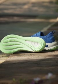 Chaussure de sport bleue avec une semelle vert vif reposant sur une surface en pierre à l'extérieur sous une lumière douce du soleil.