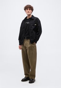 Νέος άνδρας με σγουρά μαλλιά που φοράει μαύρο σακάκι, μαύρο μπλουζάκι, μπεζ παντελόνι φαρδύ, μαύρη ζώνη και μαύρα παπούτσια, στέκεται μπροστά σε απλό φόντο.