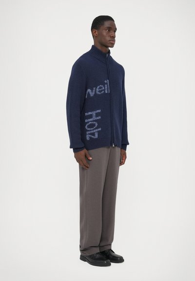 Marinblå zip-up tröja i en strukturerad stickning, med ljusblå text på framsidan. Matchad med grå byxor och svarta skor.