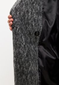 Abrigo de lana gris con un exterior texturizado, que presenta un gran botón negro y un forro de satén negro. Detalle de la costura y el ojal visible.