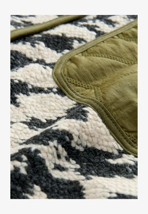 Material texturat în crem, negru și verde măslinie, prezentând o suprafață pufoasă cu o margine netedă, quiltuită în verde măslinie și detalii de cusătură.