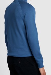 Pull-over bleu à manches longues avec col montant, en tissu doux, présentant une texture lisse et des poignets côtelés. Pantalon en velours côtelé noir visible.