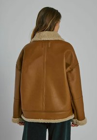 Vrouw met haar rug naar de kijker, gekleed in een bruine schapenvachtjas met een crème kleurige bont voering op de kraag, manchetten en zoom, in combinatie met donkere broek.