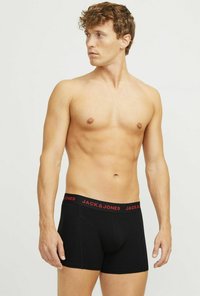 Schwarze Boxershorts mit einem roten elastischen Bund, der Text trägt. Enganliegendes Design, weicher Stoff, Oberschenkel-lange. Das Modell zeigt das Profil von der Seite.