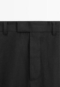 Gros plan sur la taille d'un pantalon en tissu noir avec passants pour ceinture et fermeture éclair avant dissimulée.