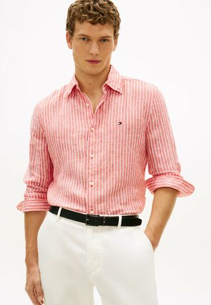 Mężczyzna w różowo-białej koszuli w pionowe paski z długim rękawem podwiniętym do łokci, białych spodniach i czarnym pasku, stojący z jedną ręką w kieszeni.