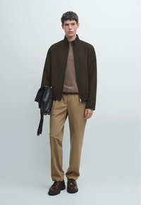 Ciemnobrązowa kurtka na zamek na beżowym swetrze i beżowych spodniach. Czarna skórzana torba trzymana w ręku. Brązowe buty z czarnymi podeszwami. Jasne tło.