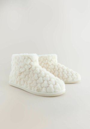 Slonovinovo biele plyšové členkové topánky s textúrovaným vzorom pom-pomu, mäkkým chlpatým materiálom a hladkou bielou podrážkou. Žiadne viditeľné kovania ani vzory.